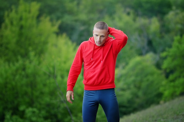 Ein schlanker sportlicher Läufer in roter Jacke mit Kapuze und schwarzer Sportleggins ruht nach dem Joggen auf einer bunten grünen Waldnatur.