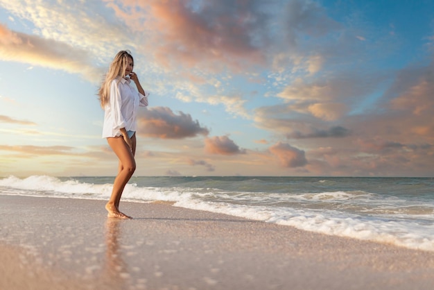 Ein schlankes junges Mädchen in einem sanften blauen Badeanzug und einem weißen Hemd geht an einem breiten Sandstrand in der Nähe des blauen Meeres mit weißen Wellen entlang