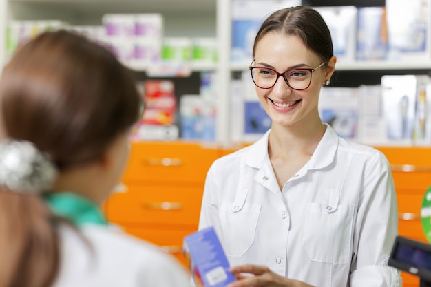 Ein schlankes Mädchen mit langen dunklen Haaren und einem weißen Kittel unterhält sich mit einem Besucher in einer Apotheke