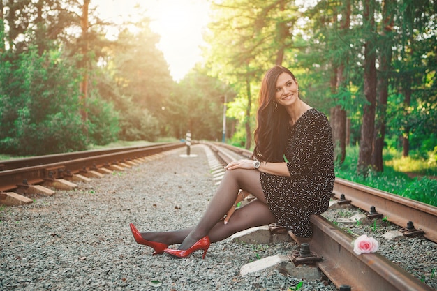 Ein schöner Brunette in einem schwarzen Kleid und roten Schuhen sitzt auf den Schienen