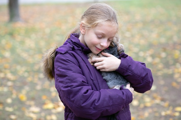 Ein schönes 9-jähriges Mädchen steht in einer lila Jacke im Park und umarmt sanft einen Welpen