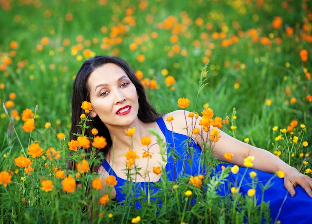 Ein schönes Altai-Mädchen in einem blauen Kleid liegt im Sommer zwischen den Blumen im Altai-Gebirge. Tourismus-Konzept.