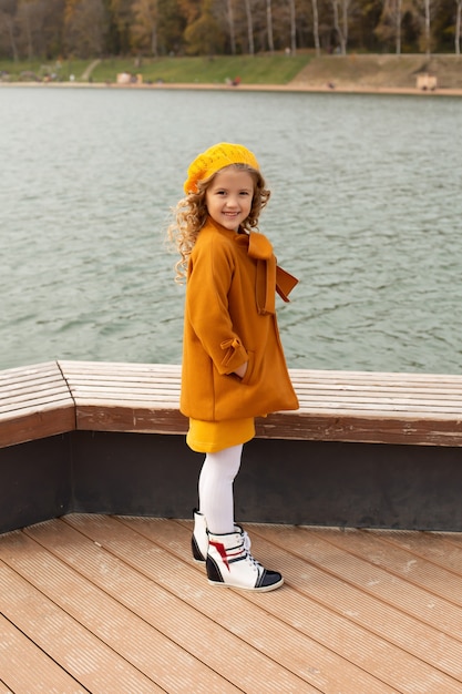 ein schönes blondes Mädchen in einem beigen Mantel in einem gelben Barett auf dem Pier des Stadtsees