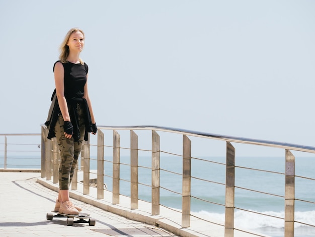 Ein schönes blondes Mädchen mit Rucksack auf Skateboard im heißen Sommertag am Meer. Wind weht ihr durchs Haar. Freiheit genießen