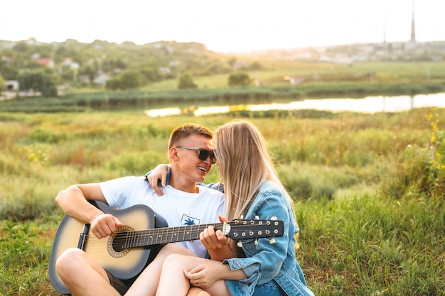 Ein schönes glückliches Paar in der Liebe mit der Sonnenbrille, welche die Gitarre spielt und bei Sonnenuntergang sich freut