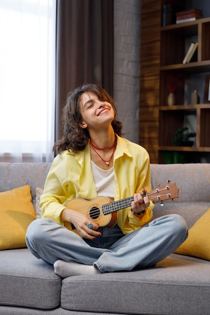 Ein schönes Hipster-Mädchen mit lockigen Haaren in einem gelben Hemd spielt und singt die Ukulele auf dem Sofa in der Wohnung Home Hobbies, die Musikinstrumente spielen