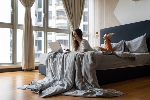 Ein schönes junges brünettes Mädchen arbeitet am Laptop, während es auf einem Bett durch ein Panoramafenster mit einer schönen Ansicht von einem hohen Stockwerk liegt. Stilvolles modernes Interieur. Ein gemütlicher Arbeitsplatz