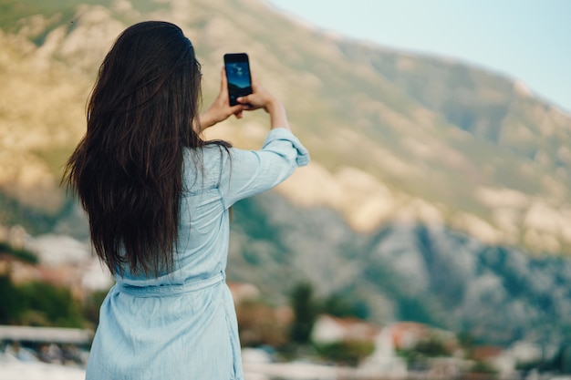 Ein schönes junges Mädchen in einem blauen Kleid, das nahe Bergen sitzt und das Telefon verwendet