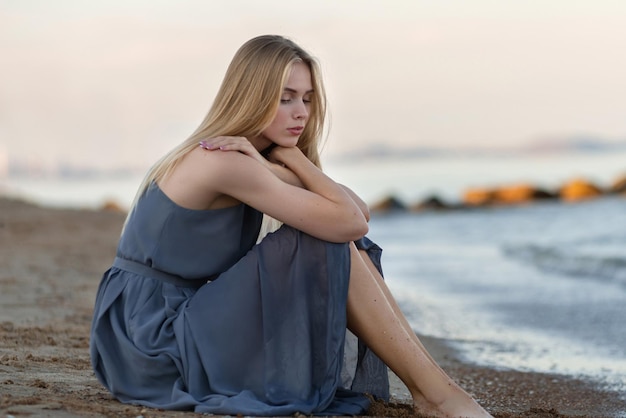 Ein schönes junges Mädchen in einem langen blauen Kleid sitzt am Meer und beobachtet den Sonnenuntergang