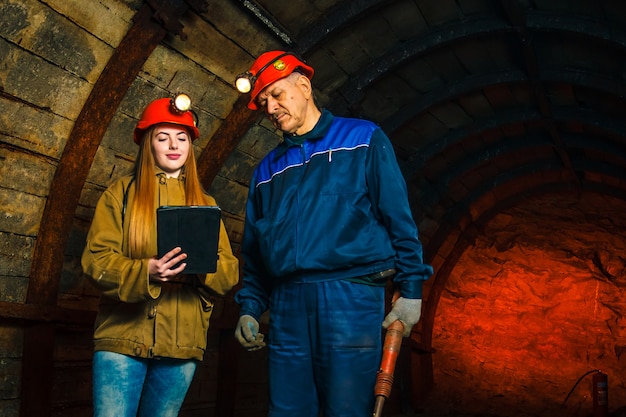 Ein schönes junges Mädchen in einem roten Helm und mit einem elektronischen Tablet in den Händen steht mit einem Bergmann in einer Kohlenmine. Businessplan Diskussion.
