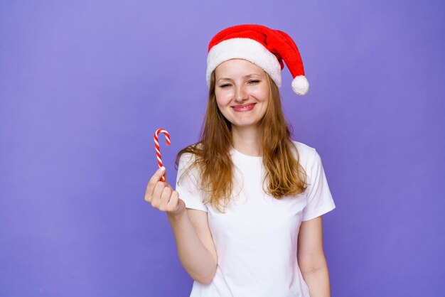 Ein schönes junges Mädchen in weißem T-Shirt und Weihnachtsmütze hält eine festliche Süßigkeit in der Hand auf einer lila Ba...