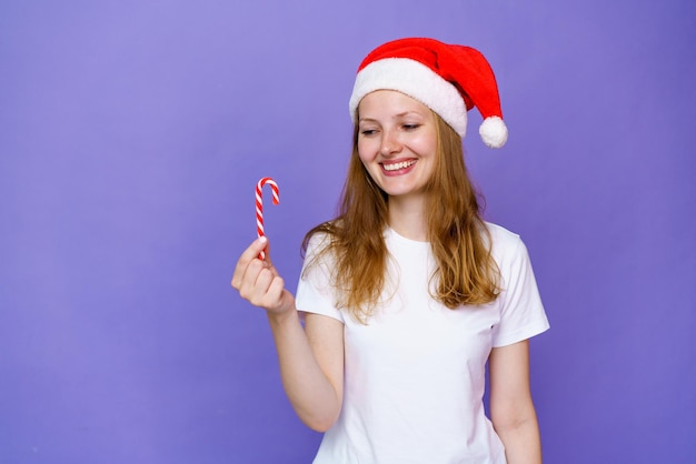 Ein schönes junges Mädchen in weißem T-Shirt und Weihnachtsmütze hält eine festliche Süßigkeit in der Hand auf einer lila Ba...