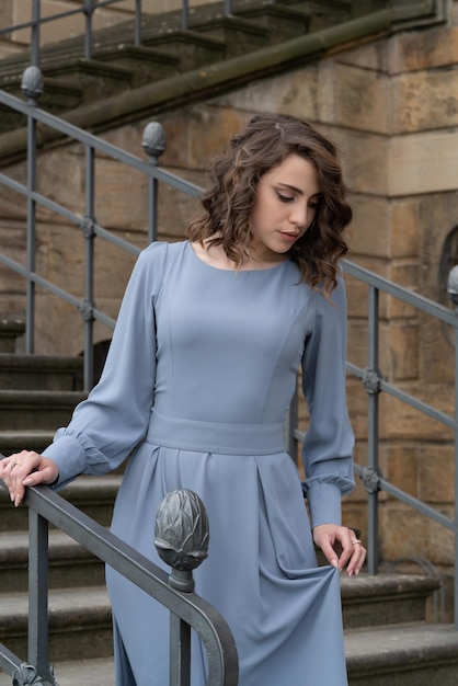Ein schönes junges Mädchen mit dunklen Haaren in einem grauen Kleid steht auf der Treppe des Schlosses und blickt nach unten Weiblichkeit Attraktivität