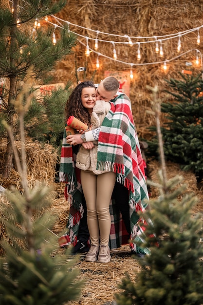 Ein schönes junges Paar in weißen Kleidern sitzt neben einem Weihnachtsbaum im Garten. Glücklicher Mann und Frau, Romantik, Weihnachtsfeier, Spaß, Liebe.