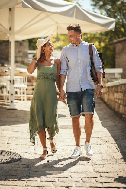 Ein schönes junges Paar vergnügt sich beim Spaziergang durch eine mediterrane Stadt. Sie genießen den sonnigen Sommertag, halten sich an der Hand und lächeln.