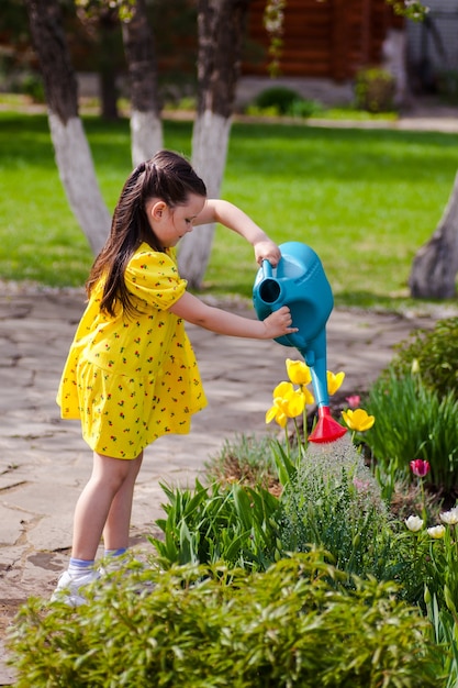 Ein schönes Kind gießt Blumen aus einer blauen Gießkanne im Garten und hilft den Eltern, sich um die ...