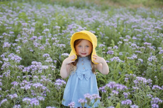 Ein schönes kleines Mädchen in einem blauen Kleid und einem gelben Hut steht auf einem Feld mit Blumen