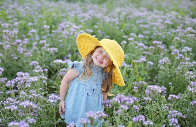 Ein schönes kleines Mädchen in einem blauen Kleid und einem gelben Hut steht auf einem Feld mit Blumen
