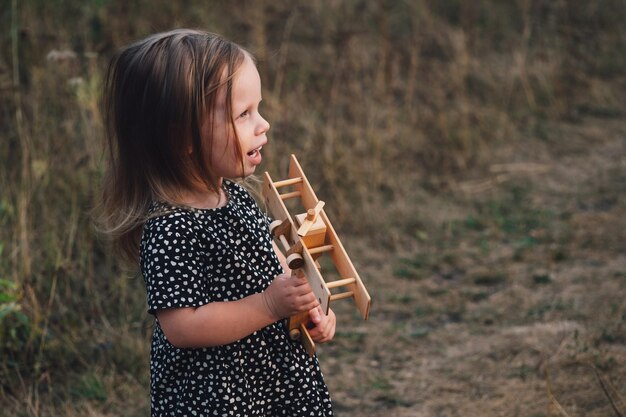 Ein schönes kleines Mädchen in einem Kleid hält ein Holzflugzeug in den Händen vor dem Hintergrund der Herbstnatur und des Sonnenuntergangs Retro-Stil Ideen für Postkarten Poster Kinderträume und Fantasien