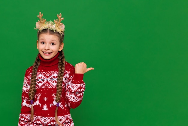 Ein schönes kleines Mädchen in einem weihnachtlichen Pullover und Schal zeigt auf einen isolierten grünen Hintergrund