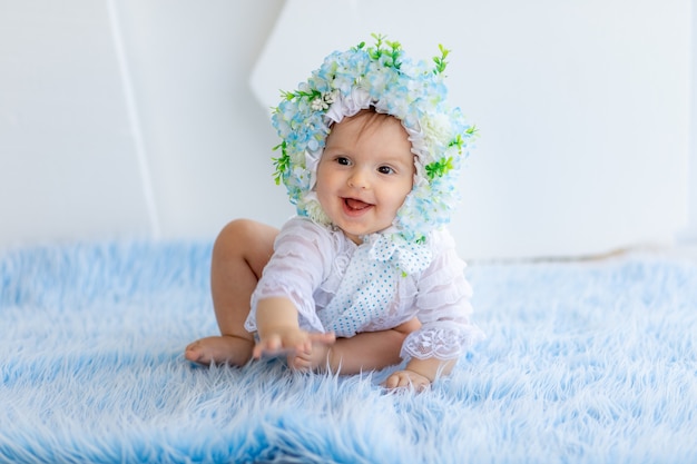 Ein schönes kleines Mädchen sitzt auf einem blauen flauschigen Teppich, trägt einen Hut aus Blumen und lächelt