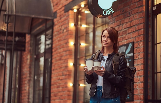 Ein schönes Mädchen, das eine Lederjacke mit einem Rucksack trägt, der draußen in der Nähe des Cafés Tassen mit Kaffee zum Mitnehmen hält.
