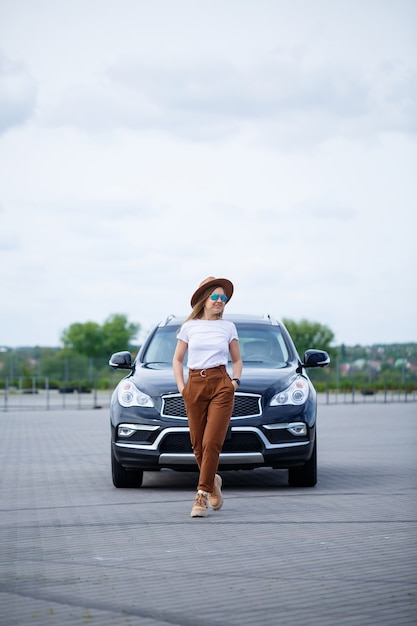 Ein schönes Mädchen europäischen Aussehens mit Brille und braunem Hut steht in der Nähe eines schwarzen Autos. Junge Frau mit Auto auf dem Parkplatz