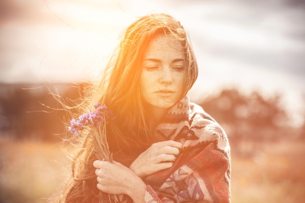 Ein schönes Mädchen genießt den Geruch von einem Strauß Kornblumen Warmer ethnischer Plaid-Sonnenuntergang Schöne rothaarige junge Dame mit Sommersprossen