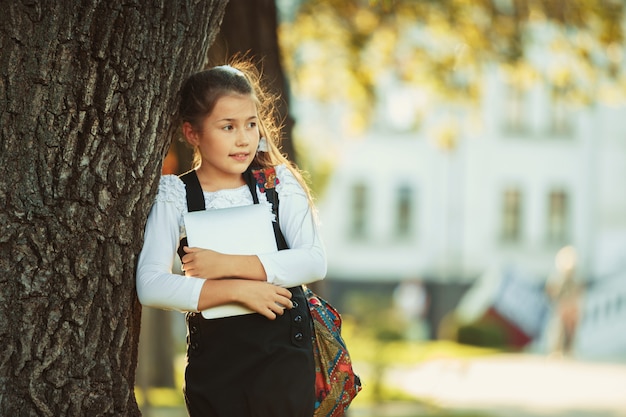 Ein schönes Mädchen im schulpflichtigen Alter steht an einem Baum und hält eine Tafel. Schulmädchen im Sommerkleid