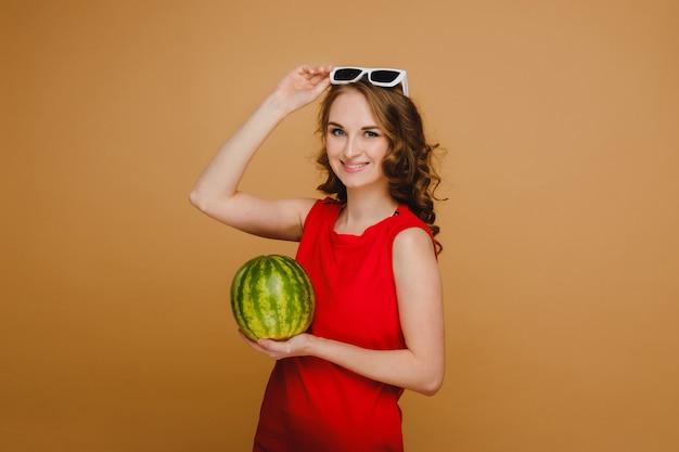 Ein schönes Mädchen in Brille und rotem Kleid hält eine Wassermelone in den Händen.