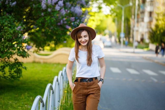 Ein schönes Mädchen in einem braunen Hut und einem weißen T-Shirt geht an einem warmen sonnigen Tag auf die Straße. Schöne junge Frau mit Hut mit einem Lächeln im Gesicht