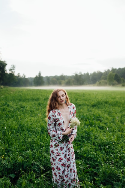 Ein schönes Mädchen in einem bunten Kleid steht am Abend auf einer Wiese vor dem Hintergrund des Nebels.