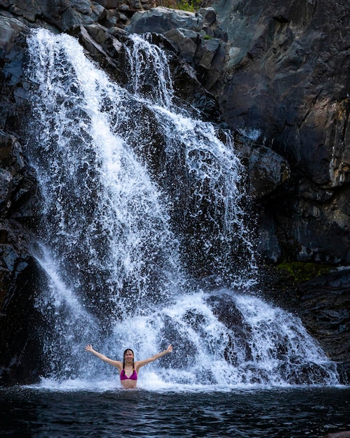 ein schönes mädchen in einem rosa bikini steht unter einem wasserfall in einem natürlichen pool, queensland, australien