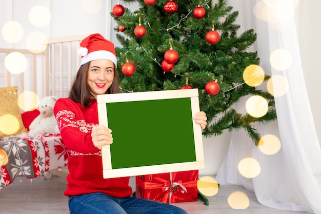 Ein schönes Mädchen in einem roten Pullover im Kinderzimmer in der Nähe des Weihnachtsbaums mit Neujahrsinterieur hält eine Tafel mit Platz für Text