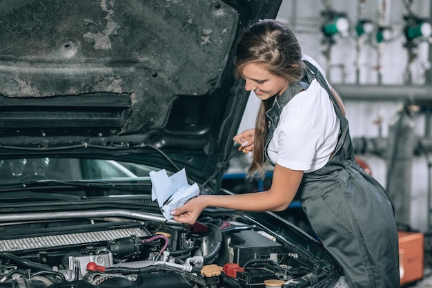 Ein schönes Mädchen in einem schwarzen Overall und einem weißen T-Shirt lächelt und überprüft den Ölstand in einem schwarzen Auto in der Garage.