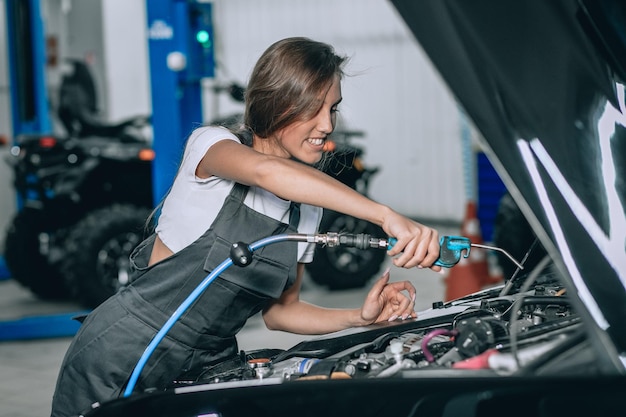 Ein schönes Mädchen in einem schwarzen Overall und einem weißen T-Shirt lächelt und überprüft den Ölstand in einem schwarzen Auto in der Garage.