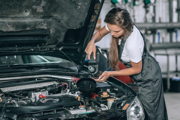 Ein schönes Mädchen in einem schwarzen Overall und einem weißen T-Shirt lächelt und überprüft den Ölstand in einem schwarzen Auto in der Garage.