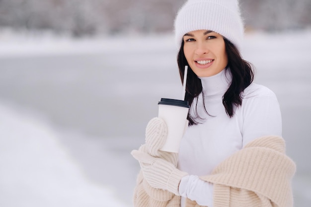 Ein schönes Mädchen in einer beigen Strickjacke und einem weißen Hut mit einem Glas Tee genießt einen schneebedeckten Damm am See