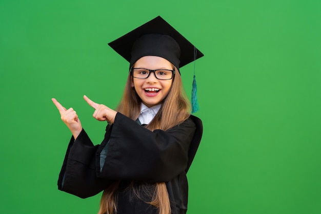 Ein schönes Mädchen in einer Schuluniform, ein Schüler zeigt mit dem Finger auf Werbung, Prüfungen am Ende des Schuljahres, ein Schulmädchen in einem Meistergewand und einer Mütze, ein Mädchen auf einem grün isolierten Hintergrund