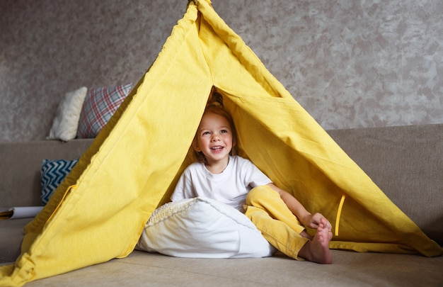 Ein schönes Mädchen in gelben Hosen und einem weißen T-Shirt spielt mit der Innenseite eines Tipis zu Hause auf der Couch. Heimspiele für Kinder