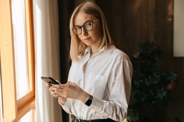 Ein schönes Mädchen in Gläsern steht am Fenster im Büro in ihren Händen mit einem Telefon, das Text tippt.