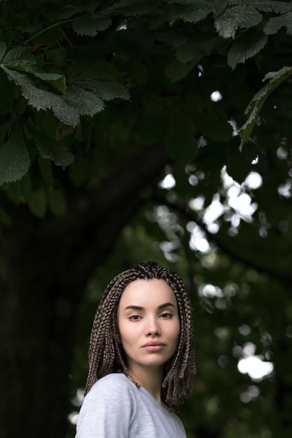 Ein schönes Mädchen mit Afro-Zöpfen blickt ernsthaft in die Kamera auf einem Hintergrund aus grünem Laub von Bäumen