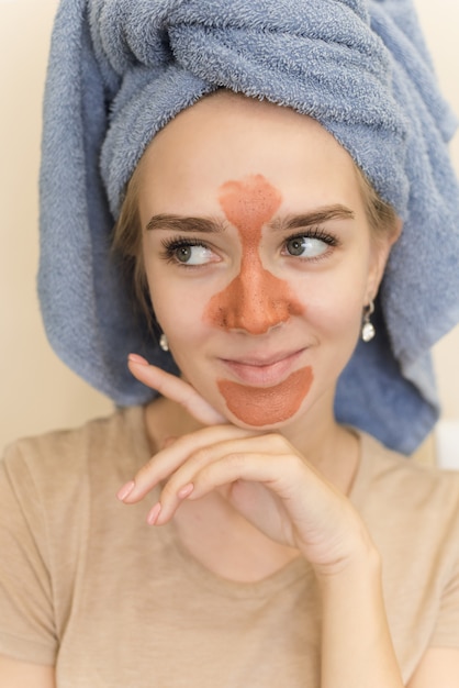 Ein schönes Mädchen mit einem Handtuch auf dem Kopf macht eine Mehrfachmaske auf der orangefarbenen T-Zone mit einem Peeling.