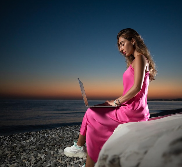 Ein schönes Mädchen mit einem Laptop in einem rosa Kleid am Meer zwischen den Felsen