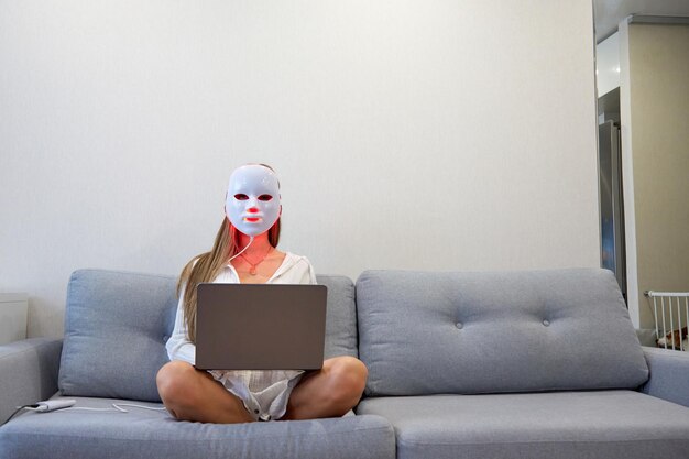 Ein schönes Mädchen mit einer LED-Maske auf dem Kopf arbeitet zu Hause an einem Laptop