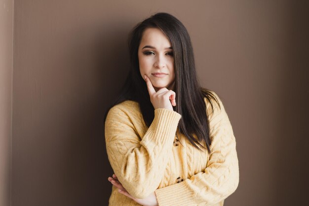Ein schönes Mädchen mit langen dunklen Haaren in einem gelben Pullover steht nachdenklich an einer schlichten braunen Wand