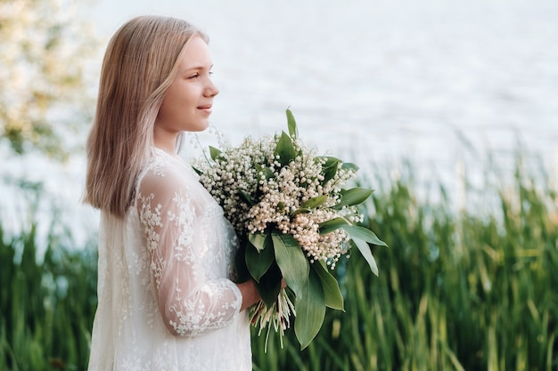 Ein schönes neunjähriges blondes Mädchen mit langen Haaren in einem langen weißen Kleid, das einen Blumenstrauß aus Maiglöckchen hält und in der Natur im Park spazieren geht.Sommer, Sonnenuntergang.