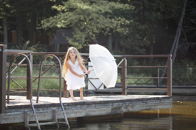 Ein schönes sechsjähriges Mädchen in einem kurzen weißen Kleid steht auf dem Mauerwerk in der Nähe des Flusses und hält einen weißen Regenschirm in den Händen