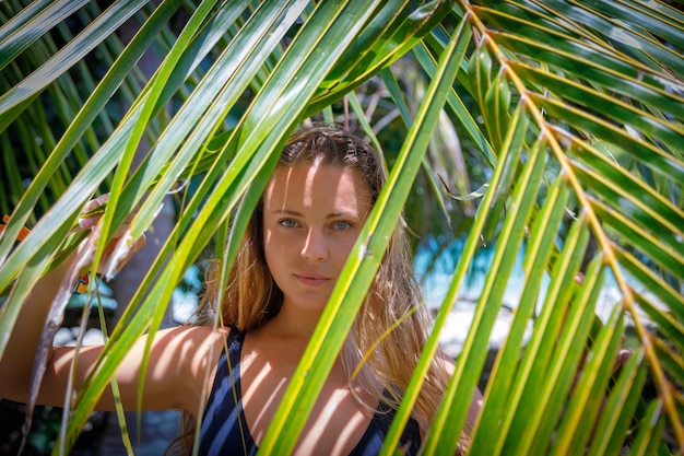 Ein schönes weißes Mädchen mit langen Haaren schaut durch ein großes Palmblatt auf die Person am Meer