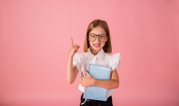 Ein Schulmädchen in Uniform und Brille hält ein blaues Notizbuch und zeigt mit dem Finger auf einen rosa Hintergrund mit einem Platz für Text